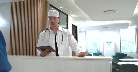 Asian male doctor greeting patient at modern clinic reception desk holding tablet