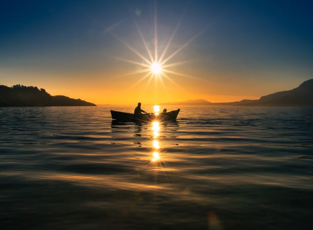 Silhouetted canoe with two people drifting on serene lake at sunrise. Smooth waters mirror the rising sun, creating a peaceful and inspiring scene. Suitable for promoting outdoor activities, travel destinations, nature conservation campaigns.