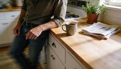 Leaning man holding coffee mug beside stacked newspaper on sunlit wooden kitchen counter