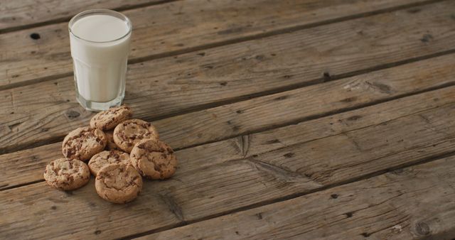Homemade chocolate chip cookies are arranged beside a glass of milk on a rustic wooden table. Perfect for use in food blogs, bakery advertisements, recipe books, or promotional materials for a cozy and inviting ambiance.