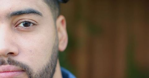 Close-up male portrait showing beard and cap, eye contact, soft blurred garden background