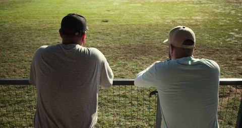 Diverse male friends wearing baseball caps leaning on railing watching sunlit sports field
