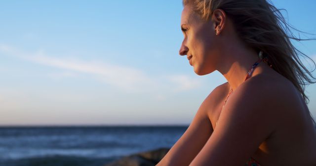 This image shows a woman enjoying a serene moment by the ocean during sunset, capturing a sense of relaxation and contemplation. Ideal for use in travel promotions, mental health articles, and advertisements emphasizing relaxation and disconnecting from hectic environments.