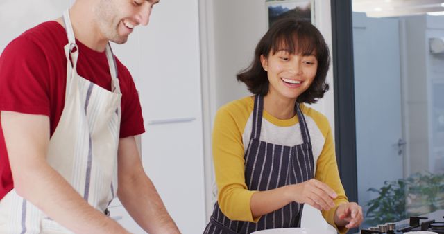 Man and woman enjoying cooking together in a modern kitchen, both smiling and wearing aprons while preparing food. Perfect for use in lifestyle blogs, cooking websites, relationship articles, or advertisements for kitchen products.