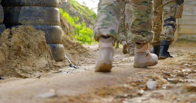 Group of soldiers walking on a dusty outdoor path, signifying military activity or training. Soldiers wear camouflage uniforms and boots. Scene includes barriers made of stacked rubber tires, contributing to the setting's rough, tactical feel. Suitable for military-themed projects, articles, or training materials, as well as editorials on defense.