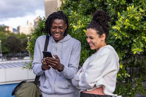 Young friends laughing while sharing smartphone on urban terrace with green foliage