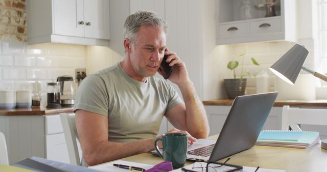 Mature man engaging in a phone call while using a laptop in a home office setting. Ideal for depicting remote work, home office setups, business communication, multitasking, and technology use in daily life.