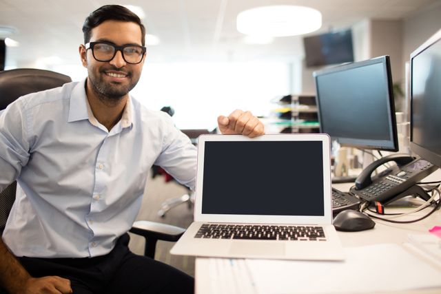 Businessman sitting at desk in modern office, smiling and showing laptop screen. Ideal for business presentations, corporate websites, technology advertisements, and professional workspace themes.