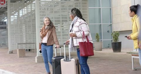 Diverse women traveling at urban curbside carrying luggage and coffee while chatting