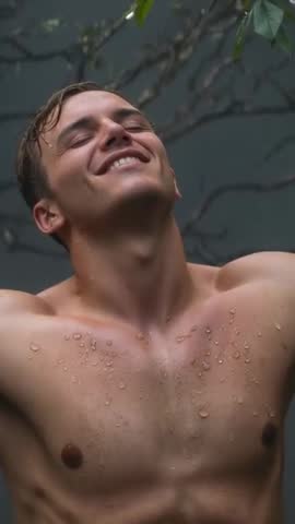 Vertical video of young man standing shirtless tilting head back under rain-soaked branches, smiling