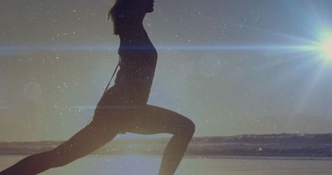 Backlit silhouette woman lunging on shoreline at sunrise with lens flare fitness lifestyle