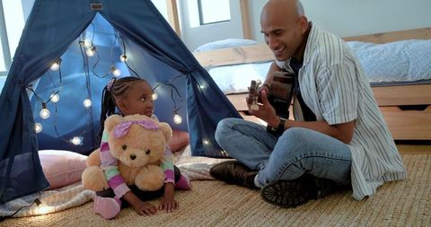 Father playing guitar with daughter in cozy bedroom teepee holding teddy bear