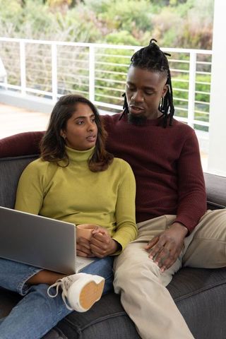 Interracial couple relaxing on balcony sofa with laptop and mug, casual sweaters, daylight