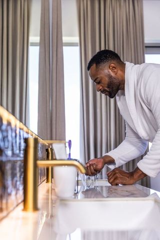 Senior african american man washing hands in robe at luxury marble sink with gold faucet