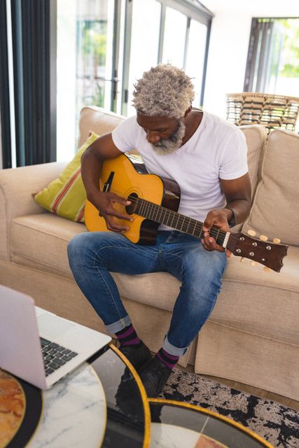 Senior African American man sitting on a sofa in a living room, practicing guitar. Ideal for use in articles or advertisements about music, hobbies, senior lifestyle, home activities, and creative pursuits.