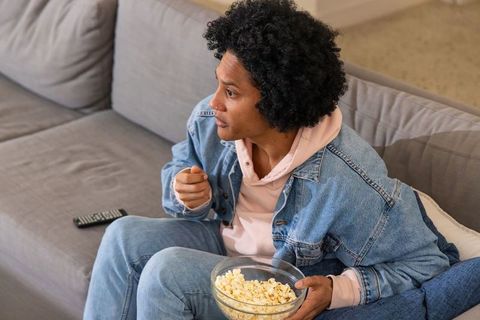African american man watching movie holding popcorn on grey couch wearing denim jacket