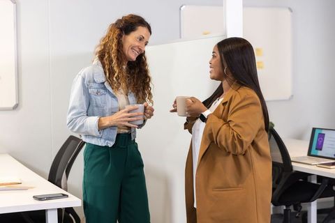 Female colleagues chatting over coffee in modern office workspace, casual team conversation