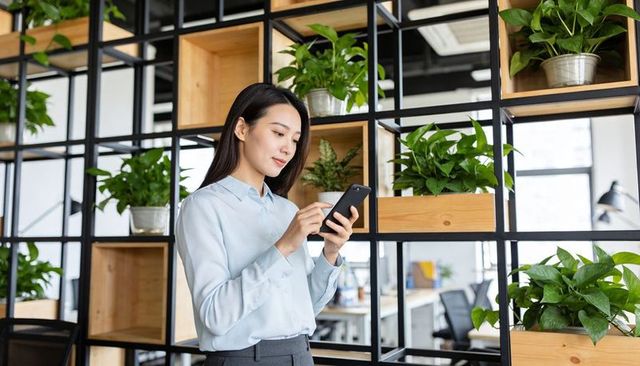 Asian woman checking phone in modern office with green plant divider and open shelving