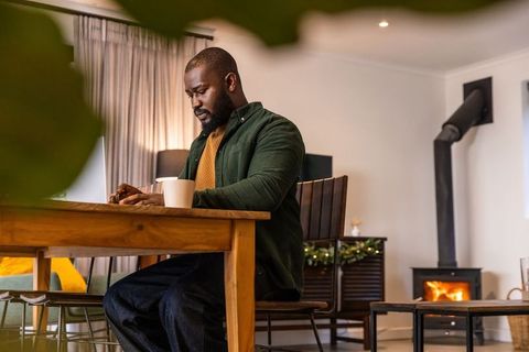 African american man sitting at wooden table using smartphone with cozy fireplace ambience