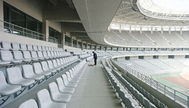 Solitary cleaner sweeping empty stadium concourse among rows of gray seats and concrete architecture