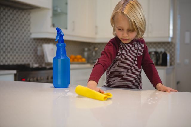 Young boy wiping kitchen counter with yellow rag, blue spray bottle nearby. Ideal for illustrating concepts of childhood chores, responsibility, and cleanliness. Suitable for parenting blogs, household cleaning products, and educational materials on teaching children domestic skills.