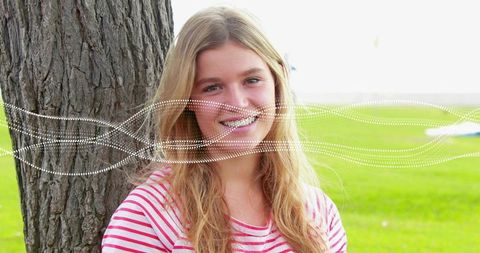Smiling young woman leaning against tree with dotted wave overlay on sunny park lawn
