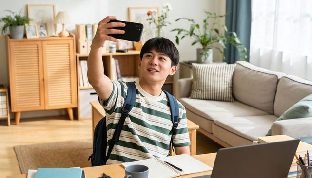 Asian male student taking selfie while studying at home with laptop, backpack, cozy workspace