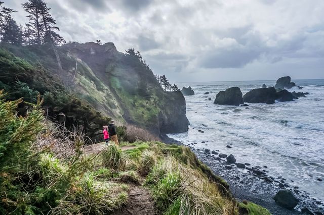Hiker exploring rugged coastal cliff overlooking ocean on a cloudy day, with crashing waves and rocky landscape. Suitable for promoting outdoor activities, nature trekking, adventure tourism, and environmental conservation.
