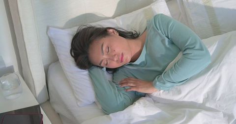 Asian woman resting peacefully in bed wearing teal top with bedside glass and red-trimmed device