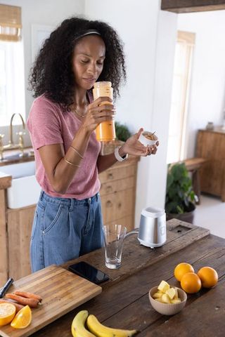 African american woman preparing orange smoothie in modern rustic kitchen with fresh fruit