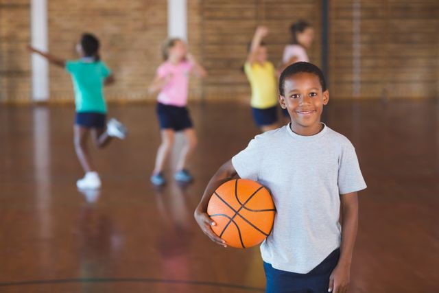 Boy standing in school gym holding basketball, smiling at camera. Other children playing in background. Ideal for educational materials, sports programs, youth activities, and fitness promotions.