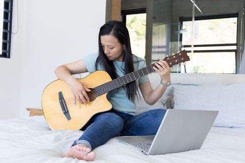 Woman playing acoustic guitar on bed, following online lessons on laptop at home, barefoot