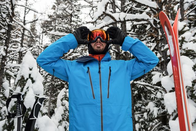 Man in blue jacket adjusting his goggles while preparing for skiing in a snowy forest. Ideal for use in winter sports promotions, outdoor adventure advertisements, and cold weather gear marketing.