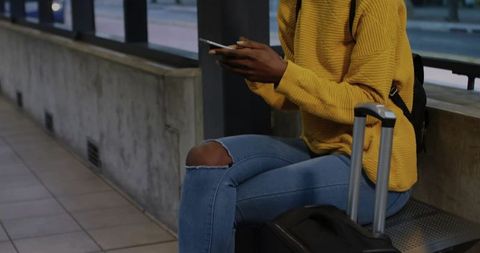 Solo traveler scrolling smartphone while waiting on urban transit platform with suitcase