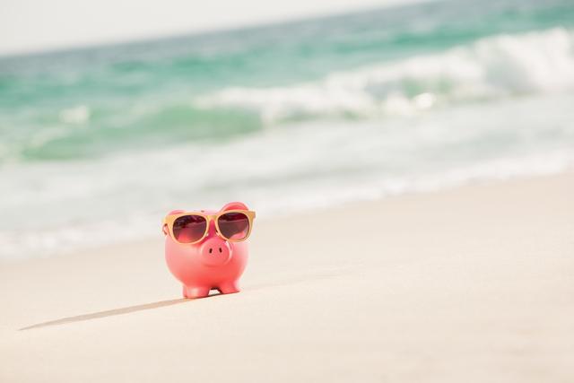 Piggy bank wearing sunglasses on beach sand with ocean waves in background. Ideal for concepts related to summer savings, vacation planning, financial goals, and fun holiday imagery. Perfect for advertisements, blog posts, and social media content promoting financial planning, travel deals, or summer activities.