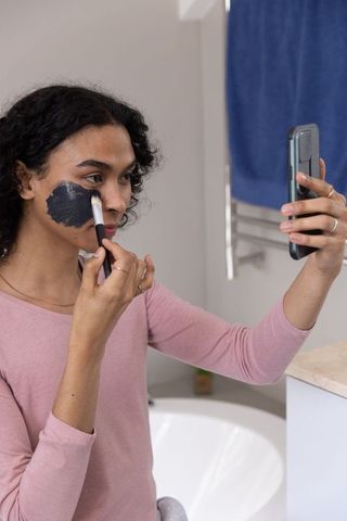 Indian non-binary applying charcoal face mask with brush while holding smartphone near tub