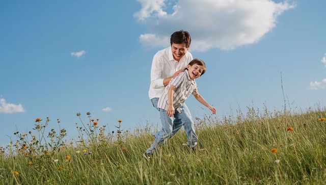 Laughing father guiding son down grassy hillside with wildflowers under blue sky