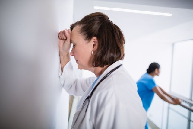 Doctor in white coat leaning against wall, appearing stressed and fatigued, with a nurse in the background. Useful for illustrating healthcare worker burnout, stress in the medical profession, and the emotional toll of working in healthcare settings.