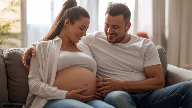Smiling expecting couple touching bare pregnant belly on sofa, intimate maternity moment