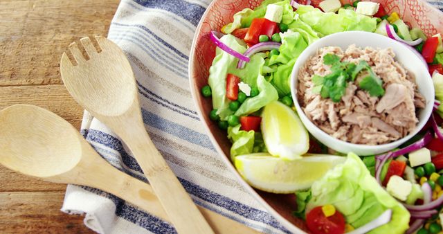 Bright, colorful salad with tuna, green peas, red peppers, onions, and lemon wedges in a bowl on wooden table. Three wooden serving utensils next to the bowl enhance rustic look. Ideal for promoting healthy eating, food blogs, recipe websites, cookbooks, and diet meal planning.