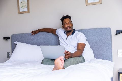 African american man using laptop on bed in minimalist bedroom for remote work and relaxation
