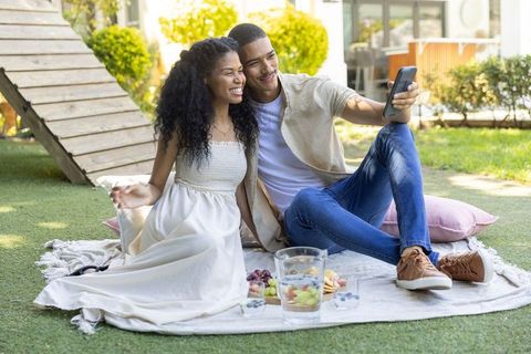 Young couple taking selfie during relaxed backyard picnic on sunny afternoon with smartphone