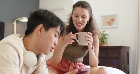 Young couple sharing smartphone on bed, woman holding coffee mug and smiling, cozy morning