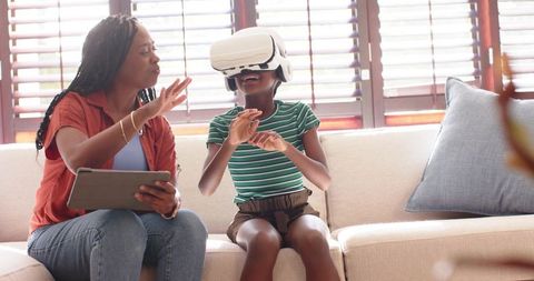 African mother guiding daughter using VR headset and tablet on sunlit living room sofa