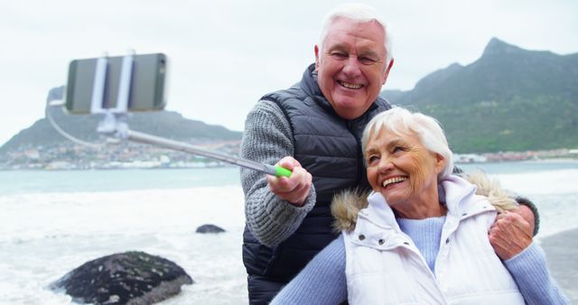 Senior couple capturing a joyful moment at the beach with a scenic mountain backdrop. Perfect for promoting travel, leisure, and adventure activities aimed at older adults. Ideal for retirement lifestyle content, vacation packages, and advertisements focusing on senior well-being and happiness.