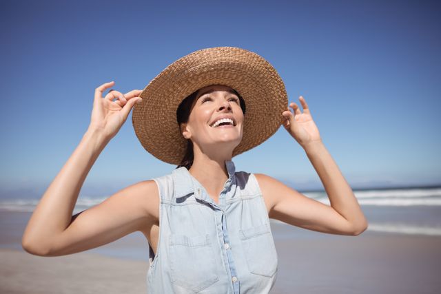 Woman wearing a sun hat, smiling and enjoying a sunny day at the beach. Ideal for use in travel brochures, summer vacation promotions, lifestyle blogs, and advertisements focusing on outdoor activities and relaxation.