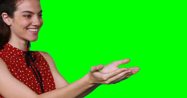 This stock photo depicts a cheerful woman in a red polka dot dress, stretching out her hands as if presenting or showing something. Ideal for use in advertisements, presentations, and educational videos where you can superimpose additional images or text on the green screen background. Perfect for marketing, business promotions, or instructional content.