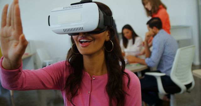 A woman is smiling and exploring virtual reality while wearing a VR headset at an office. Colleagues are collaborating on another project in the background. Perfect for use in articles, blog posts, and marketing materials showcasing modern technology in work environments and promoting innovative workplace solutions.