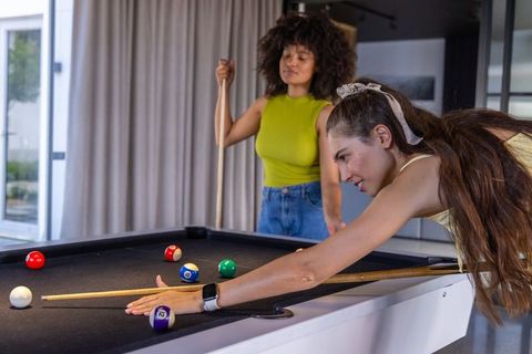 Female friends leaning and aiming cue while playing pool on black-felt table in modern lounge