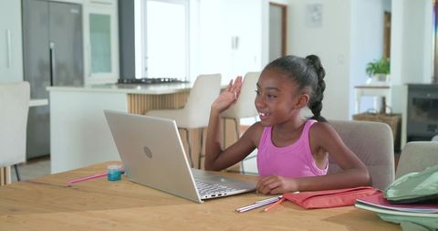 African american girl waving and smiling during remote learning on laptop at home study table